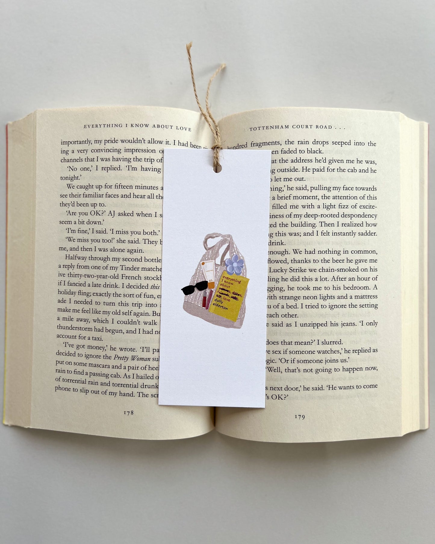 Open book with a bookmark featuring a bag, a book called everything i know about love, rhode lip balm, sunglasses and key charm on a light gray background