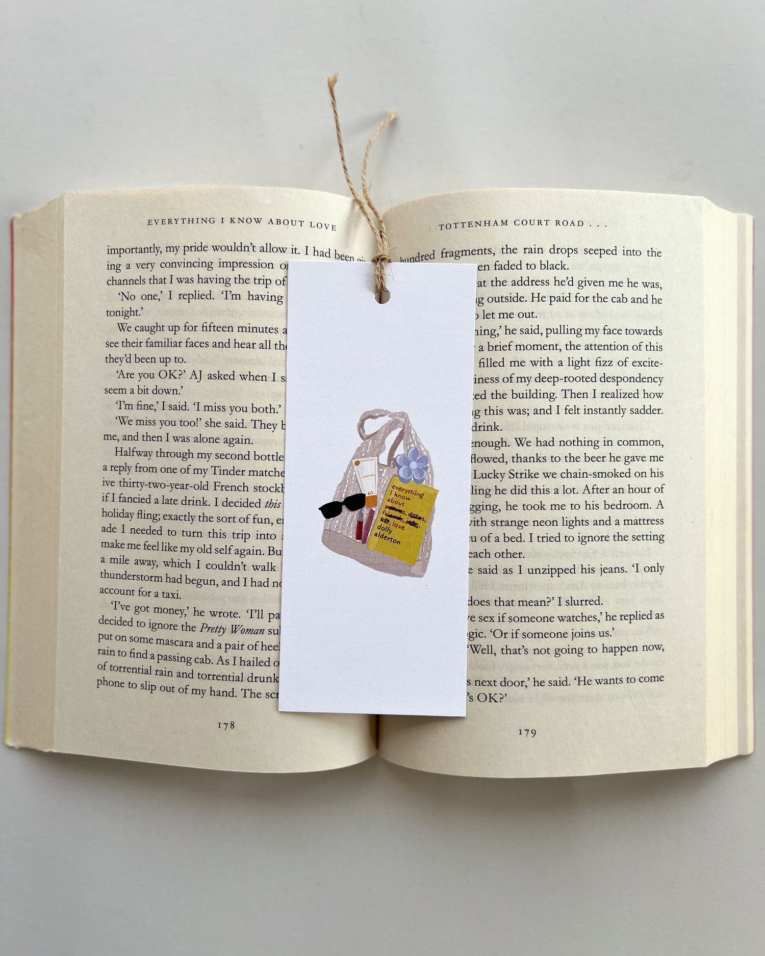 Open book with a bookmark featuring a bag, a book called everything i know about love, rhode lip balm, sunglasses and key charm on a light gray background