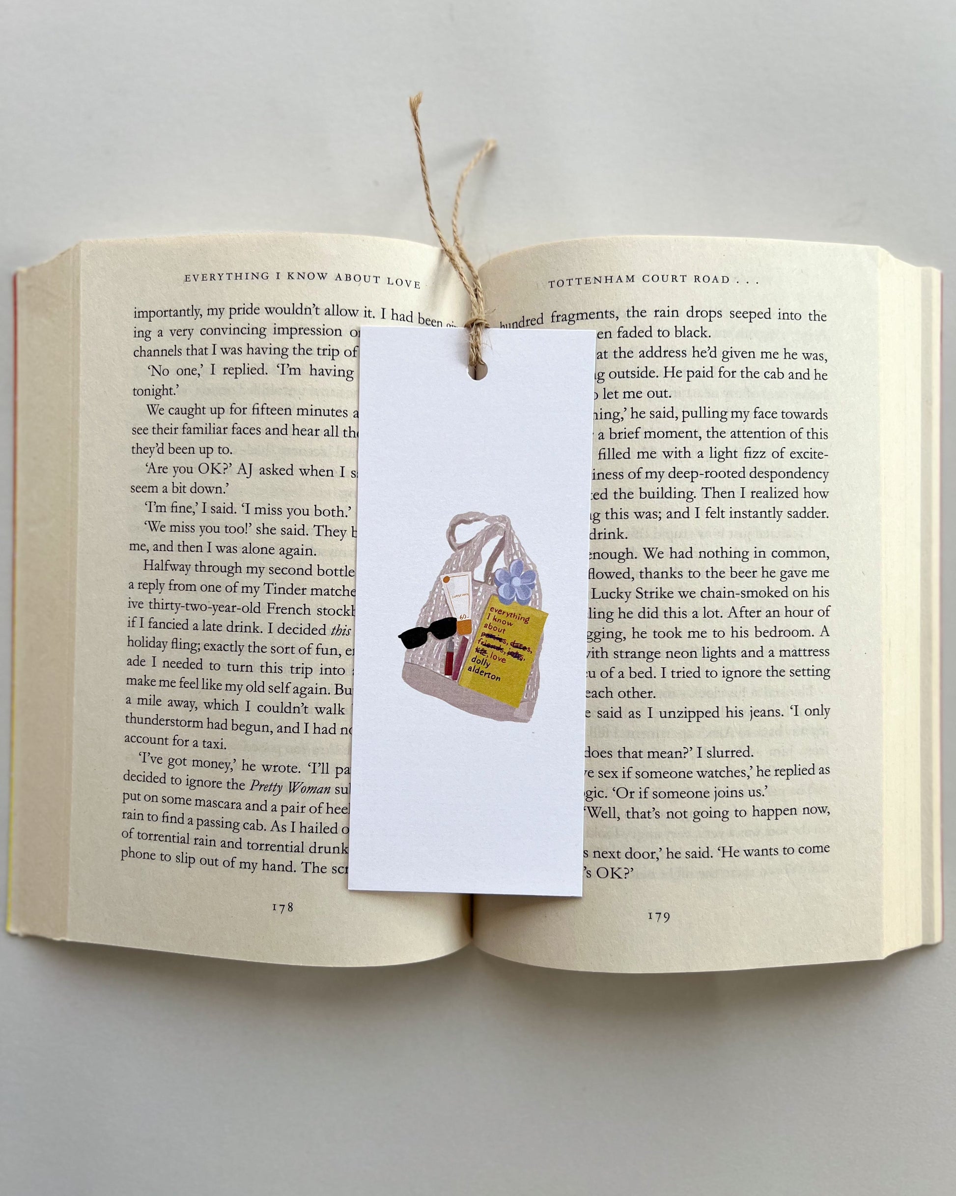 Open book with a bookmark featuring a bag, a book called everything i know about love, rhode lip balm, sunglasses and key charm on a light gray background
