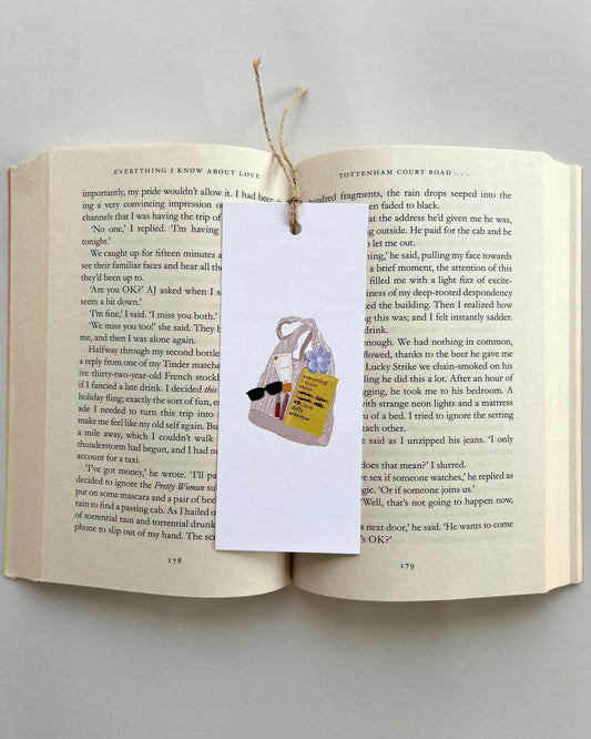 Open book with a bookmark featuring a bag, a book called everything i know about love, rhode lip balm, sunglasses and key charm on a light gray background