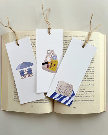 Three decorative bookmarks on an open book with illustrations of summer bags and beach chairs for a beach day.